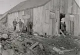 A black and white photo of three men standing beside a blacksmith shop. Carriage wheels and plywood surround them on the ground.