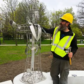 Colour photo of artist Geordie Lishman posing beside his metal art installation called Gather. Geordie is wearing a safety vest as he glances up happily at his sculpture.