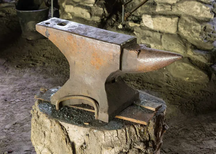 Colour photo of a rusted London pattern anvil inside of a blacksmith shop. One end is square edged and the other end has a horned shaped extension.  