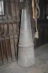 Colour photo of a big rusted floor cone mandrel. It is a tall and wide cone made from metal. It sits on the floor of a blacksmith shop, with other metal tools surrounding it.