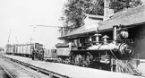 A black and white photo of an early locomotive parked at a station. Three men stand on the platform beside the locomotive.