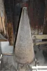 Colour photo of a big rusted floor cone mandrel. The cone is made from metal and is tall and wide. The cone mandrel sits on the floor of a blacksmith shop.