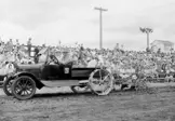 A black and white photo of an early automobile used as farming equipment. A large crowd of people sit on the bleachers behind the automobile.