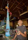 Colour photo of artist Bill Lishman admiring his colourful metal art sculpture. A tall metal cone sits in the middle of the sculpture and various small figures riding cars and bicycles made out of metal surround it.