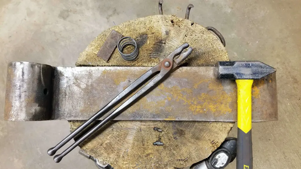 Colour photo of wolf jaw tongs placed on a metal hinge next to a yellow handled hammer. The jaws of the tongs are jagged. The metal hinge rests on a tree stump. 